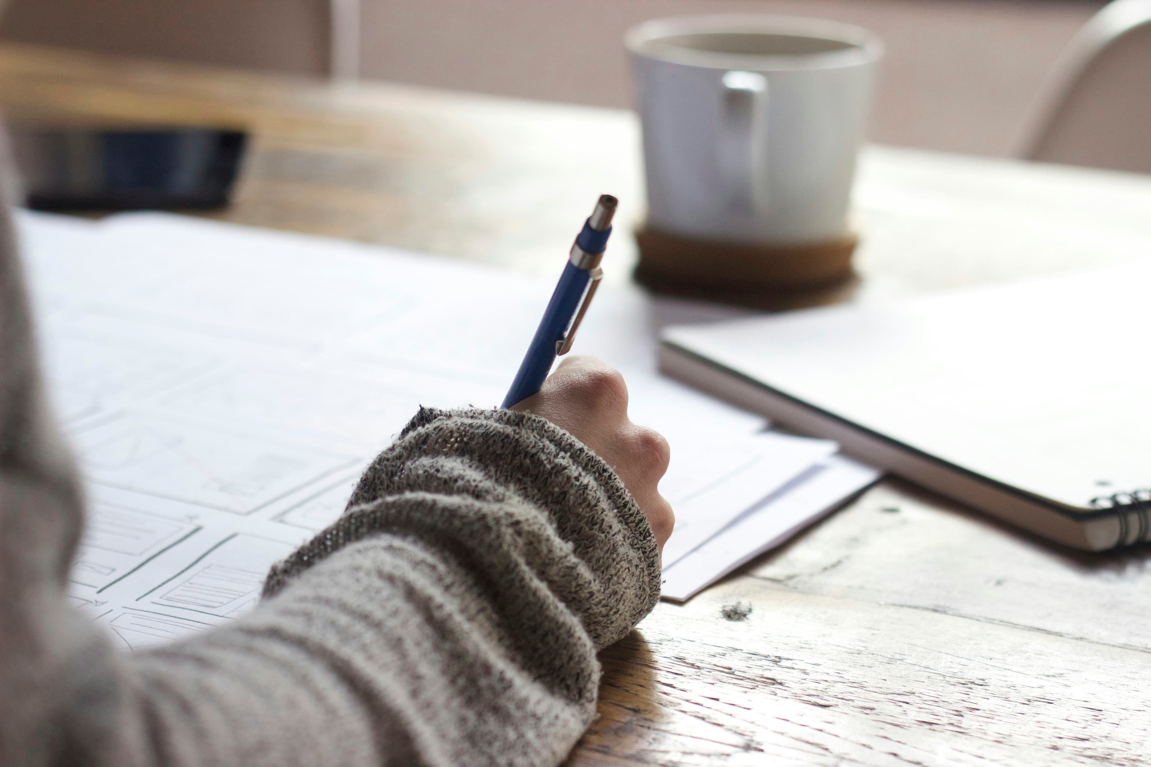 A person writing at a wooden table next to a mug