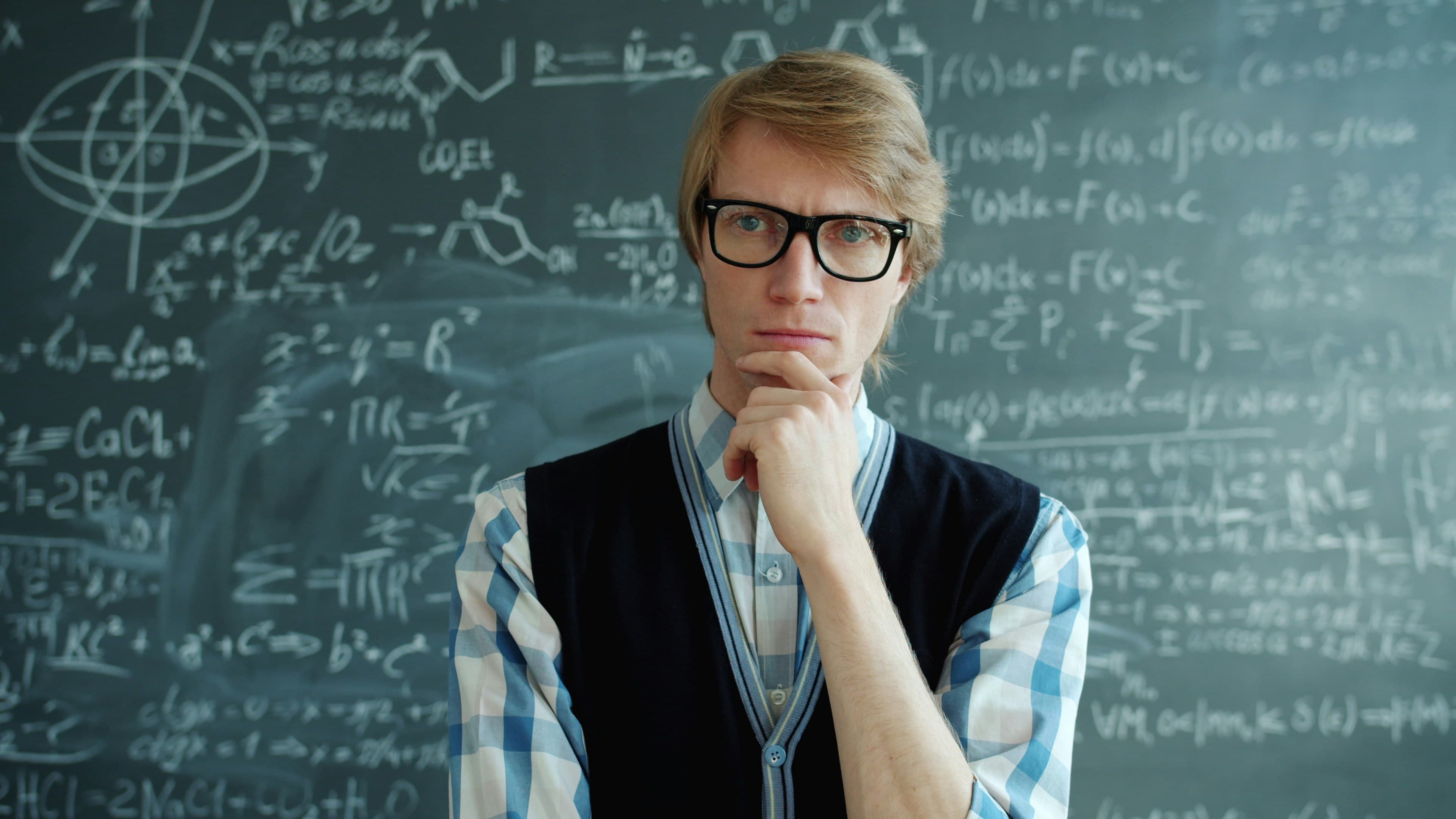 A man with glasses thinking in front of a chalkboard